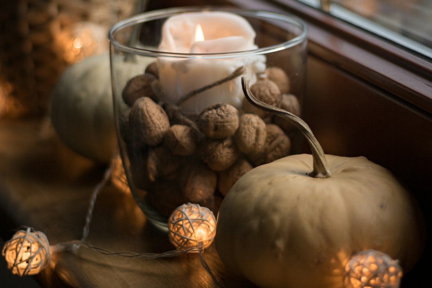 Candle in a glass filled with walnuts on a wood windowsill, pale pumpkins and glowing woven ball string lights around it, a basket with a silvery plant behind