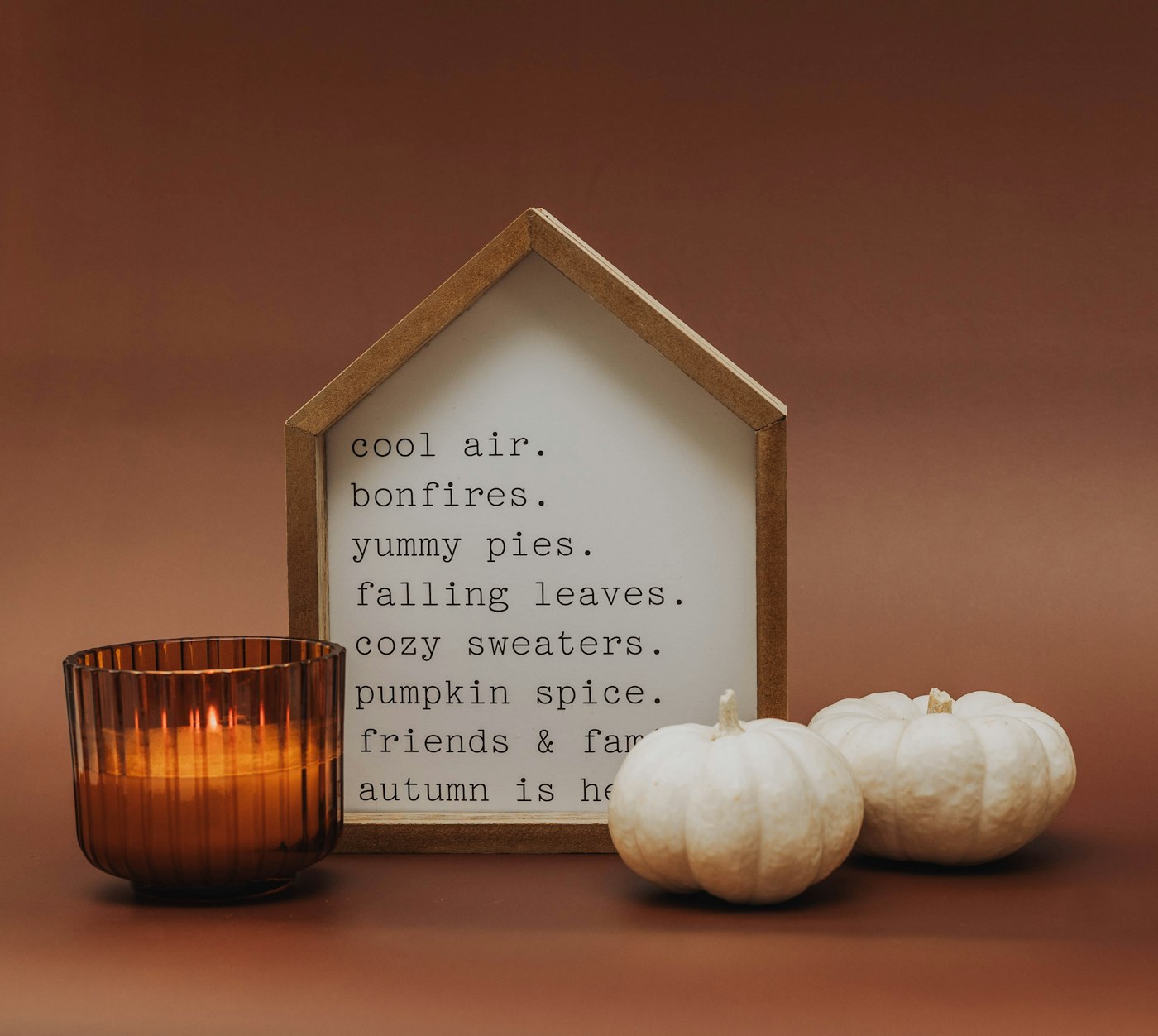 An amber ribbed glass candle, a little house-shaped fall sign and two small white pumpkins on a rust background