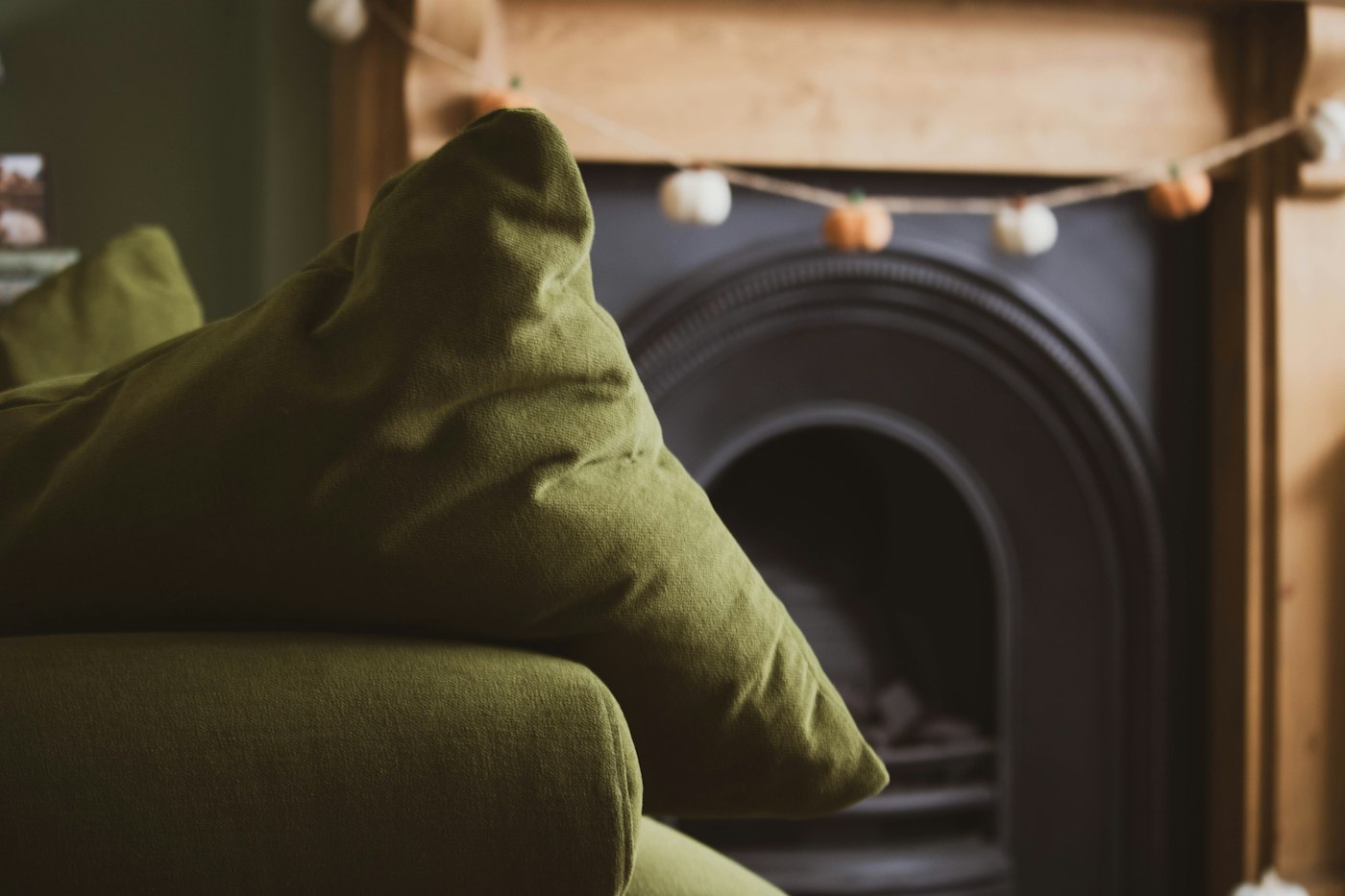 A moss green velvet cushion on a sofa, with a tiny felt pumpkin garland across a wood mantel behind it