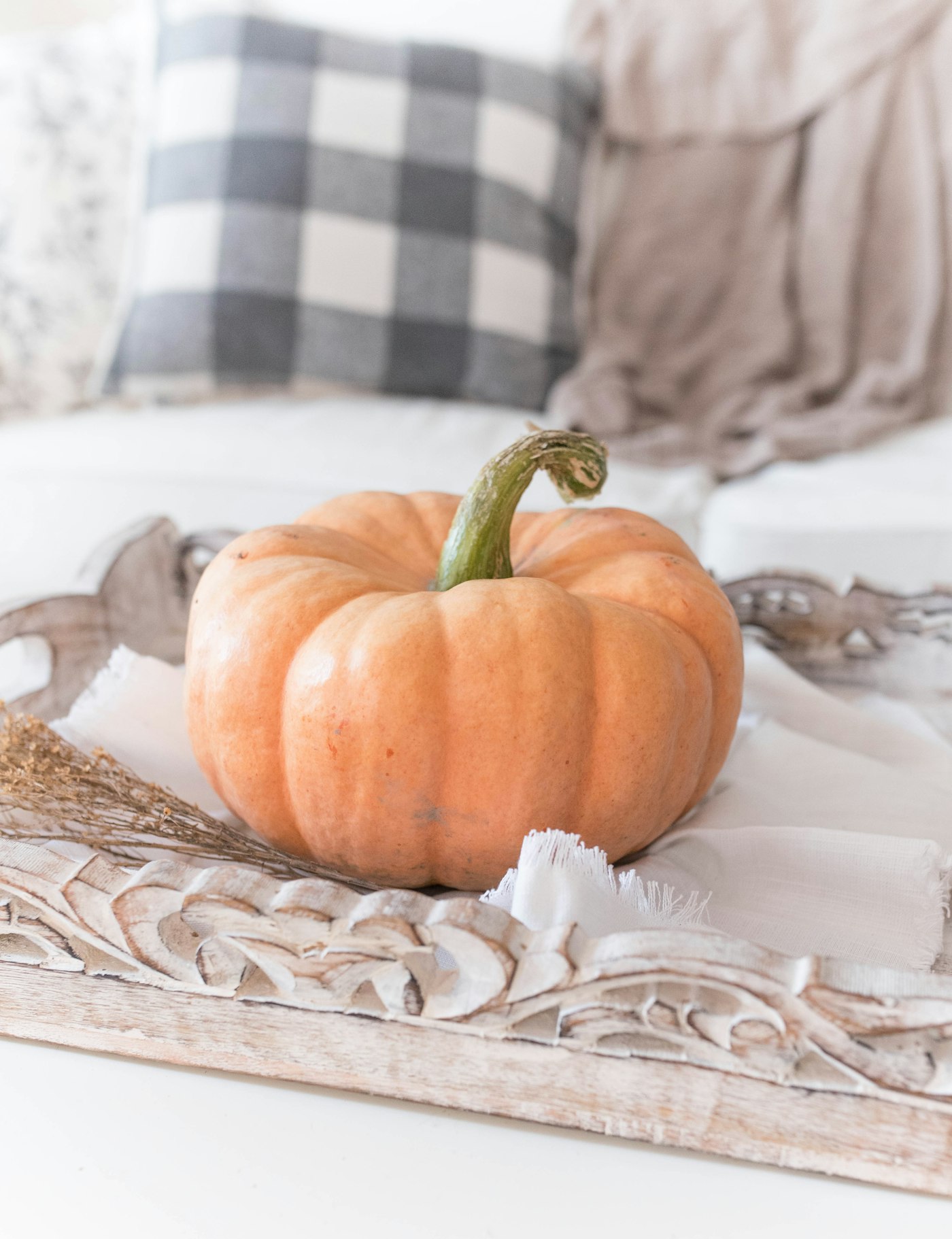 A pale peach pumpkin with a curly green stem on a carved whitewashed wood tray with frayed linen and dried flowers, a gray plaid pillow and a soft throw behind