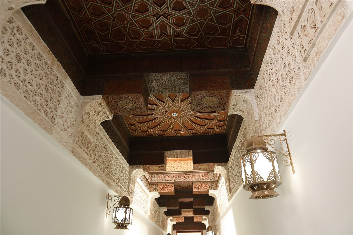 Carved and painted cedar ceiling above a white plaster hallway with brass lanterns in Morocco